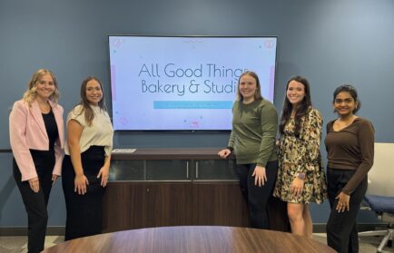 Five people standing in front of a screen displaying "All Good Things Bakery & Studio" in an office setting.