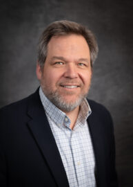 A man with gray hair and a beard is smiling. He is wearing a dark blazer over a blue and white checkered shirt, set against a dark gray background.