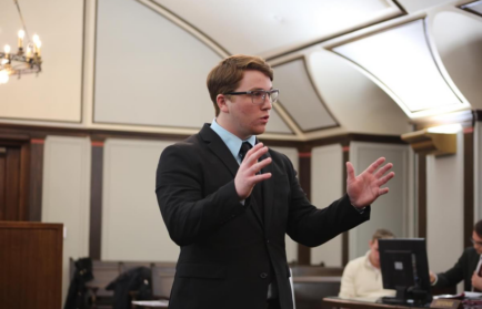 A man in a suit and glasses is speaking passionately, gesturing with both hands, in a courtroom or formal meeting room. Another person is seated and looking at a computer in the background.