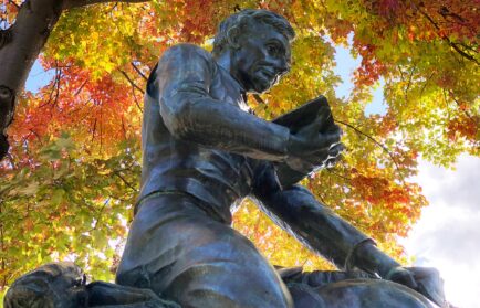 A bronze statue of a person reading a book, set against a backdrop of vivid autumn leaves on a tree.