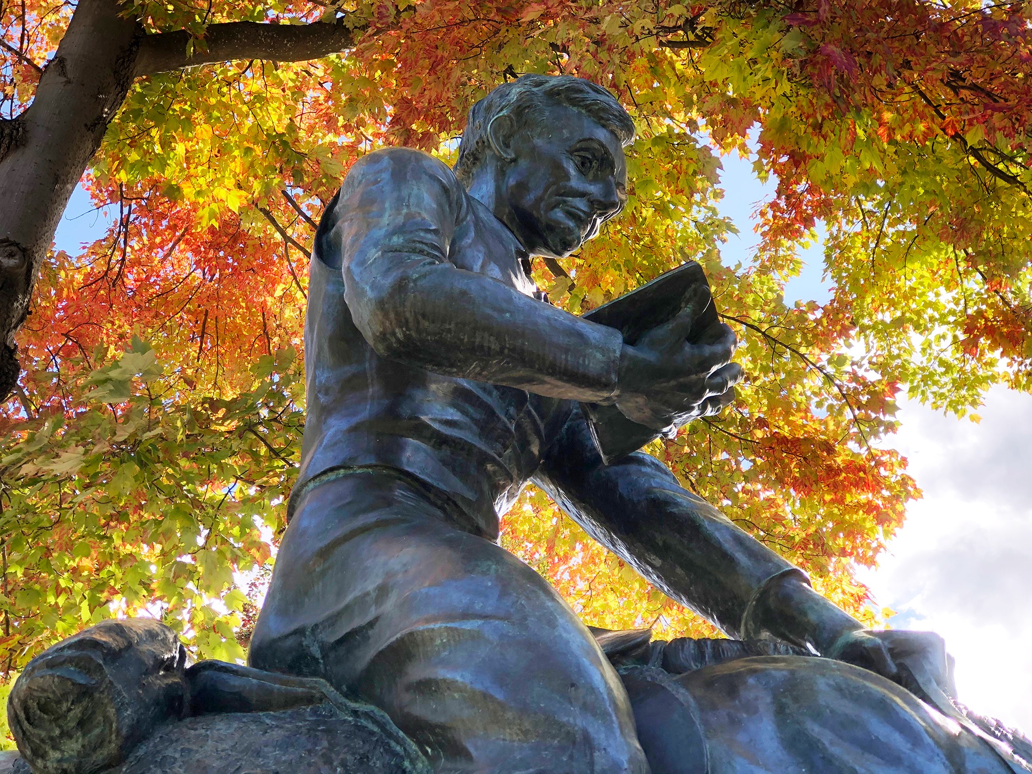 A bronze statue of a person reading a book, set against a backdrop of vivid autumn leaves on a tree.