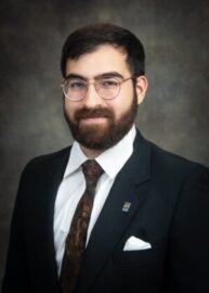 A bearded man with glasses wearing a dark suit, white dress shirt, patterned tie, and a pin on his lapel posed against a dark gray background.