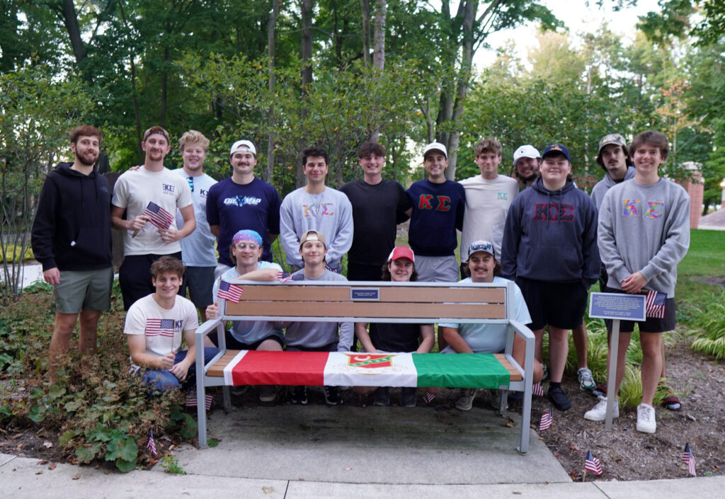 A group of 17 young men stand and sit around a bench outdoors, with a colorful banner and small American flags placed around them. Trees and foliage are in the background.
