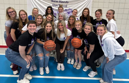 On a gym floor, a group of people confidently pose with basketballs, standing proudly in front of a banner proclaiming "These Hands Don’t Hurt," symbolizing empowerment and unity.