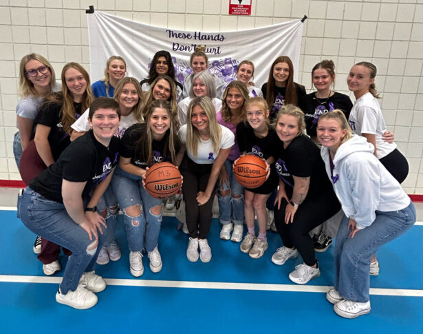 On a gym floor, a group of people confidently pose with basketballs, standing proudly in front of a banner proclaiming "These Hands Don’t Hurt," symbolizing empowerment and unity.