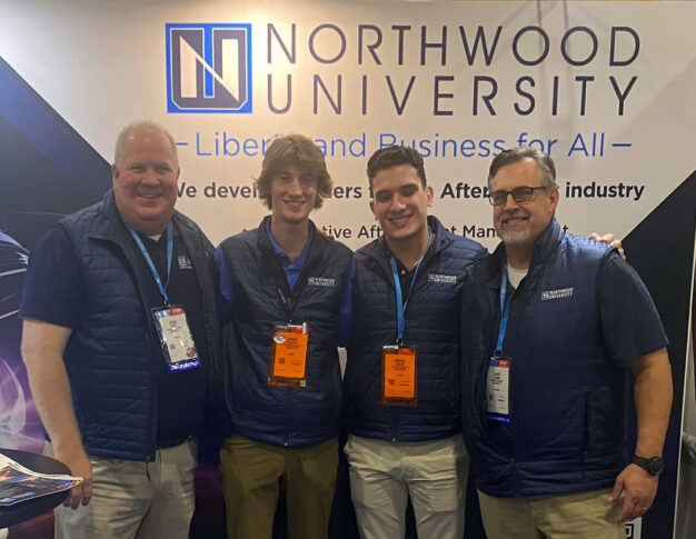 Four men in matching Northwood University vests stand together, smiling proudly, like a well-executed auto draft strategy, in front of a university banner.