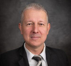 A man in a suit and tie, with short gray hair, stands against a dark background, embodying the spirit of free enterprise.