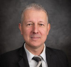 A man in a suit and tie, with short gray hair, stands against a dark background, embodying the spirit of free enterprise.