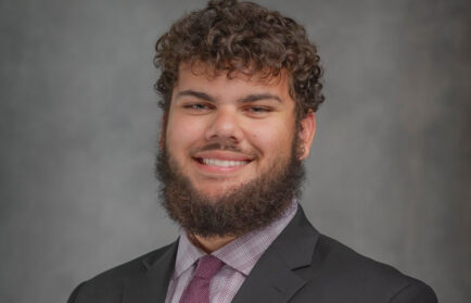 A man with curly hair and a beard, wearing a black suit, checkered shirt, and purple tie, embodies the spirit of Free Enterprise as he smiles confidently in front of a gray background.