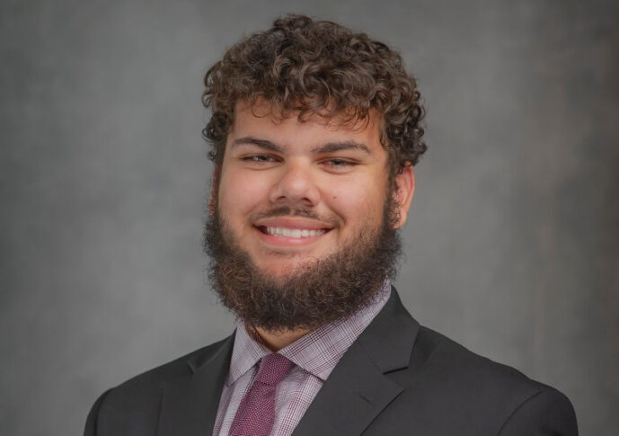 A man with curly hair and a beard, wearing a black suit, checkered shirt, and purple tie, embodies the spirit of Free Enterprise as he smiles confidently in front of a gray background.