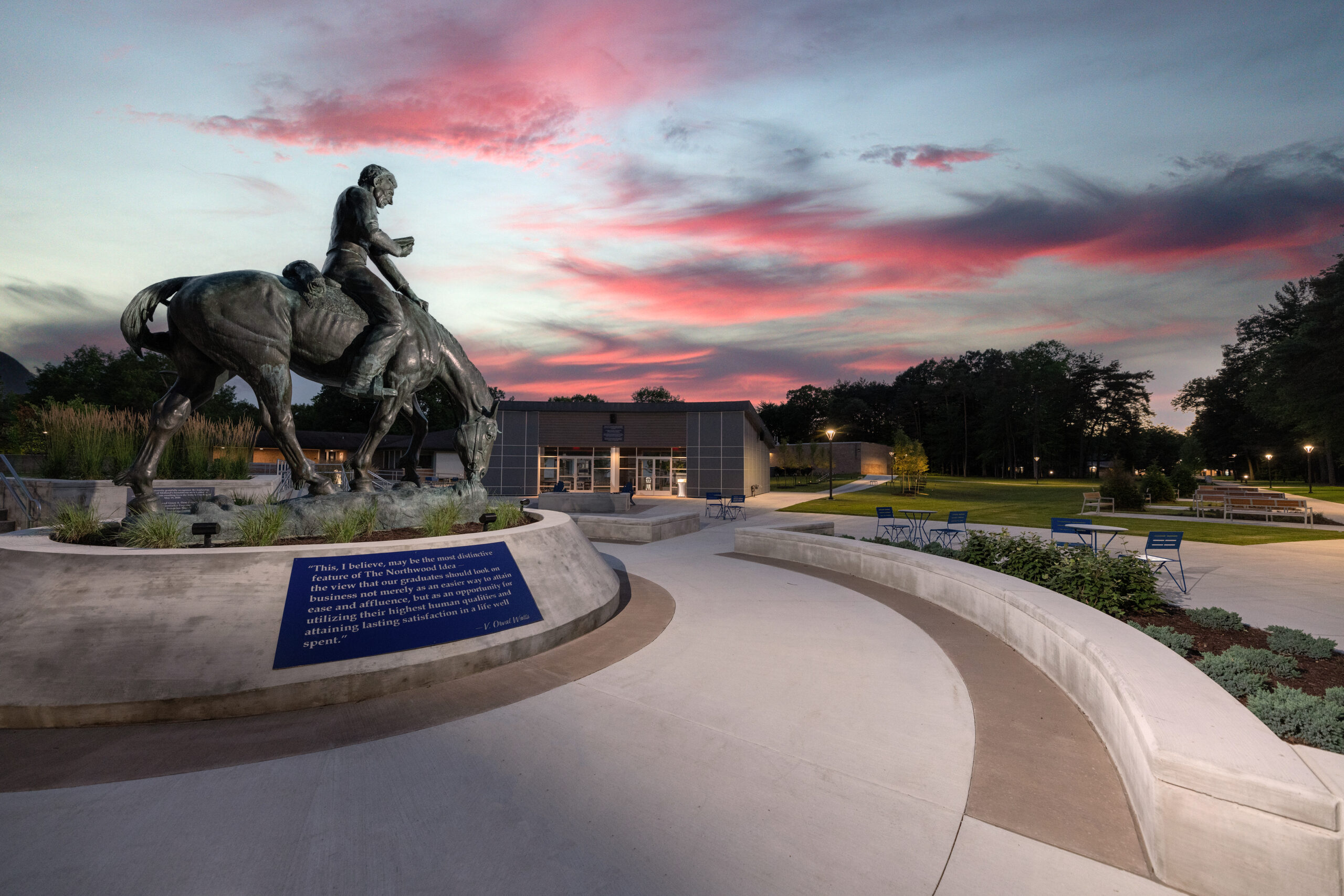 A statue of a man on horseback stands in a plaza with a blue plaque, silhouetted against the vibrant sunset sky. A modern building and seating area provide a contemporary backdrop, enhancing the blend of history and urban life.