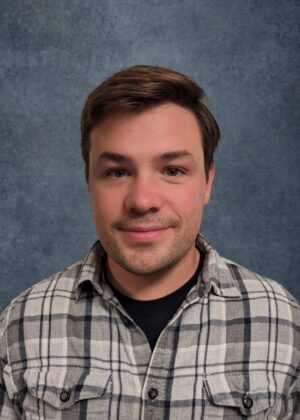 A person with short brown hair wearing a plaid shirt stands in front of a textured gray background.