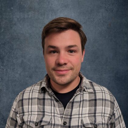 A person with short brown hair wearing a plaid shirt stands in front of a textured gray background.