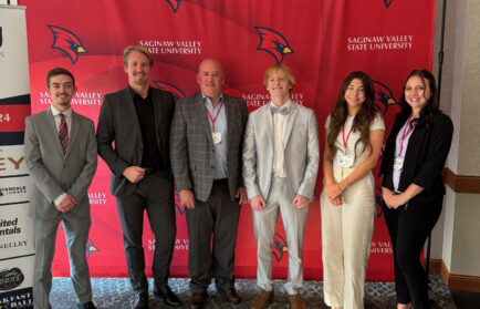 Six people stand elegantly in front of a bold red Saginaw Valley State University banner, their formal attire reflecting sophistication and poise.