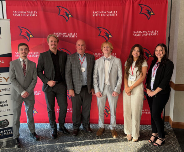 Six people stand elegantly in front of a bold red Saginaw Valley State University banner, their formal attire reflecting sophistication and poise.