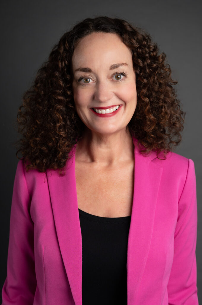 A woman with curly hair smiles while wearing a pink blazer over a black top, perhaps one of the 2024 Education Awardees, posing confidently against a dark gray background.