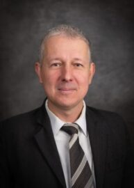 The sharp-dressed man in a suit and tie stands confidently against a dark, textured background, his poised presence creating an air of sophistication.