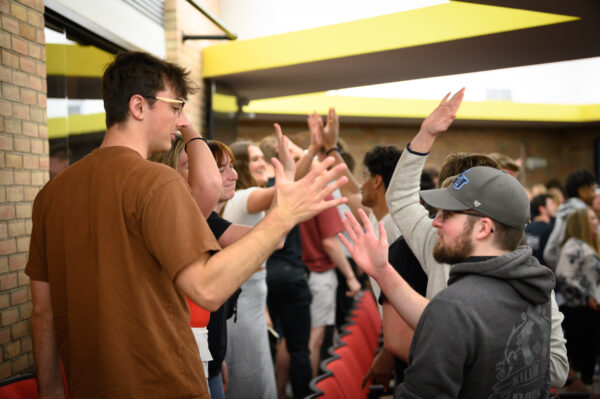 A group of admitted students in a room engage in a high-five activity while standing near rows of red chairs.