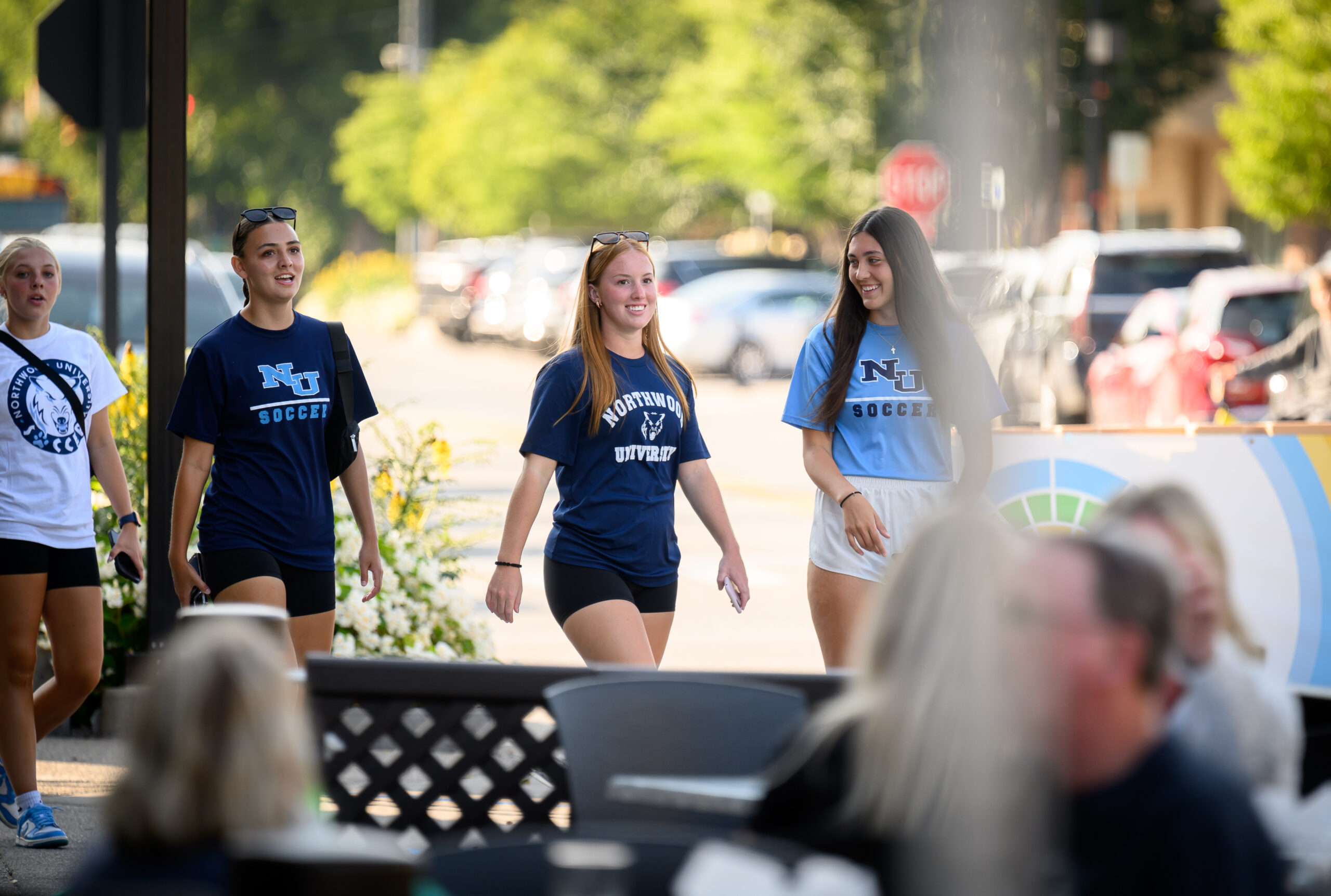 Four young women, who look like newly admitted students, are walking outdoors in blue and black t-shirts with "NU Soccer" and "Western United" logos. Trees and cars form the backdrop of their carefree stroll.