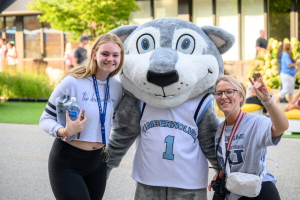 Two admitted students pose with a person in a Timberwolves mascot costume outdoors. One holds a camera and makes a peace sign.