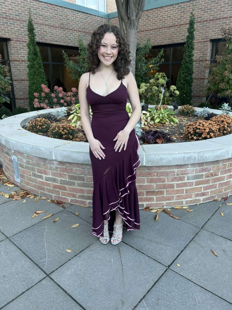 A person in a dark purple dress stands in front of a brick planter with flowers and greenery, evoking the elegance of a fashion showcase.