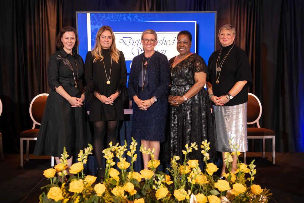 Five women stand on stage, dressed formally, against a backdrop displaying a sign about Auto Draft. Yellow flowers are arranged elegantly in the front.