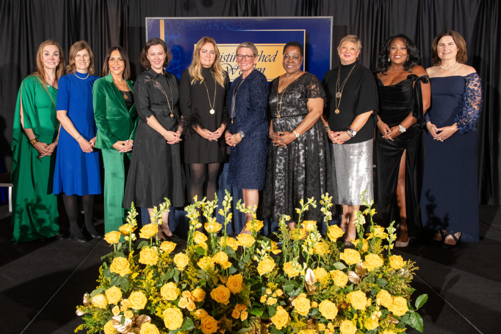 A group of eleven women in formal attire stands on stage in front of a floral arrangement and a sign reading "Auto Draft: Distinguished.