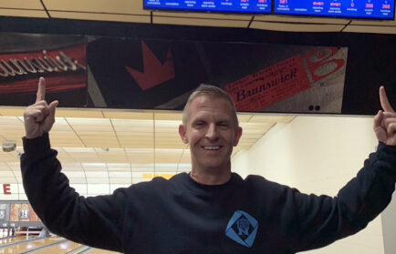 A man wearing a black sweatshirt stands in the bowling alley, pointing at the scoreboard displays above the lanes, as if strategizing his next strike with precision akin to an auto draft.