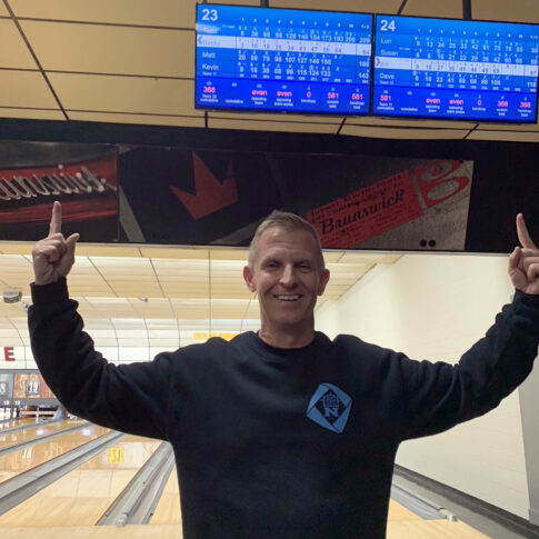 A man wearing a black sweatshirt stands in the bowling alley, pointing at the scoreboard displays above the lanes, as if strategizing his next strike with precision akin to an auto draft.