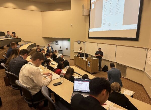 People seated in a high school lecture hall, intently listening to a presentation. A projected screen at the front displays essential details. Desks are adorned with notebooks and drinks, as students rise to the challenge of grasping new concepts.