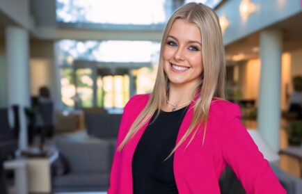 In an office setting, a person with long blonde hair and a bright pink blazer smiles, reminiscent of the vibrant energy typical at the Auto Show scheduled for Sept. 19-21 in 2025.