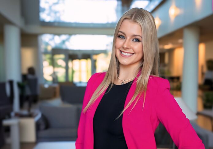 In an office setting, a person with long blonde hair and a bright pink blazer smiles, reminiscent of the vibrant energy typical at the Auto Show scheduled for Sept. 19-21 in 2025.