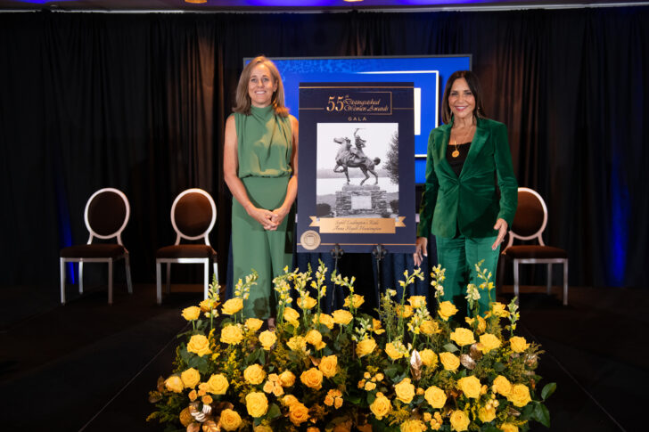 Two women in green outfits stand beside a framed poster at an event, while vibrant yellow and white flowers are arranged in the foreground, capturing the attention of attendees like an auto draft capturing ideas.