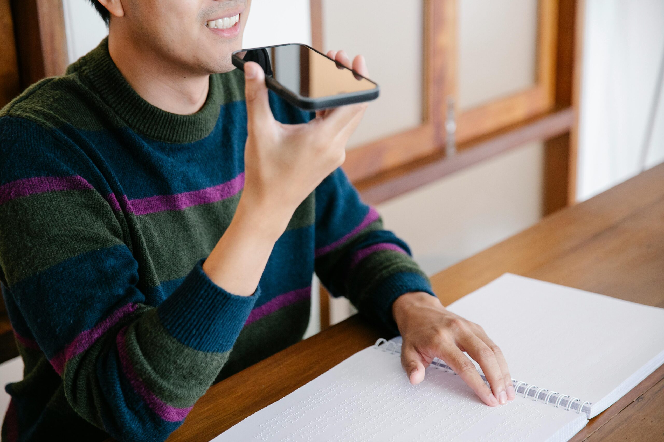 An admitted student holds a smartphone near their mouth, using voice input while resting a hand on a book on the table.