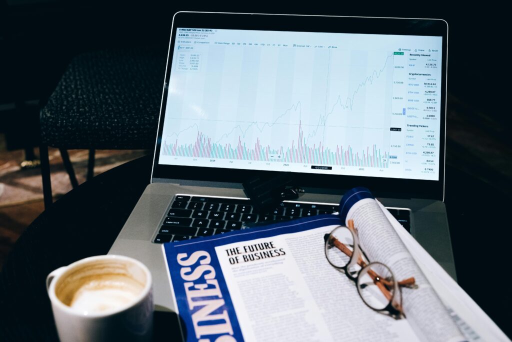 An open laptop displaying a stock market chart sits on a table alongside a magazine titled "Business," eyeglasses, and a cup of coffee, creating the perfect Emporos setting for those who thrive in the world of trading and finance.
