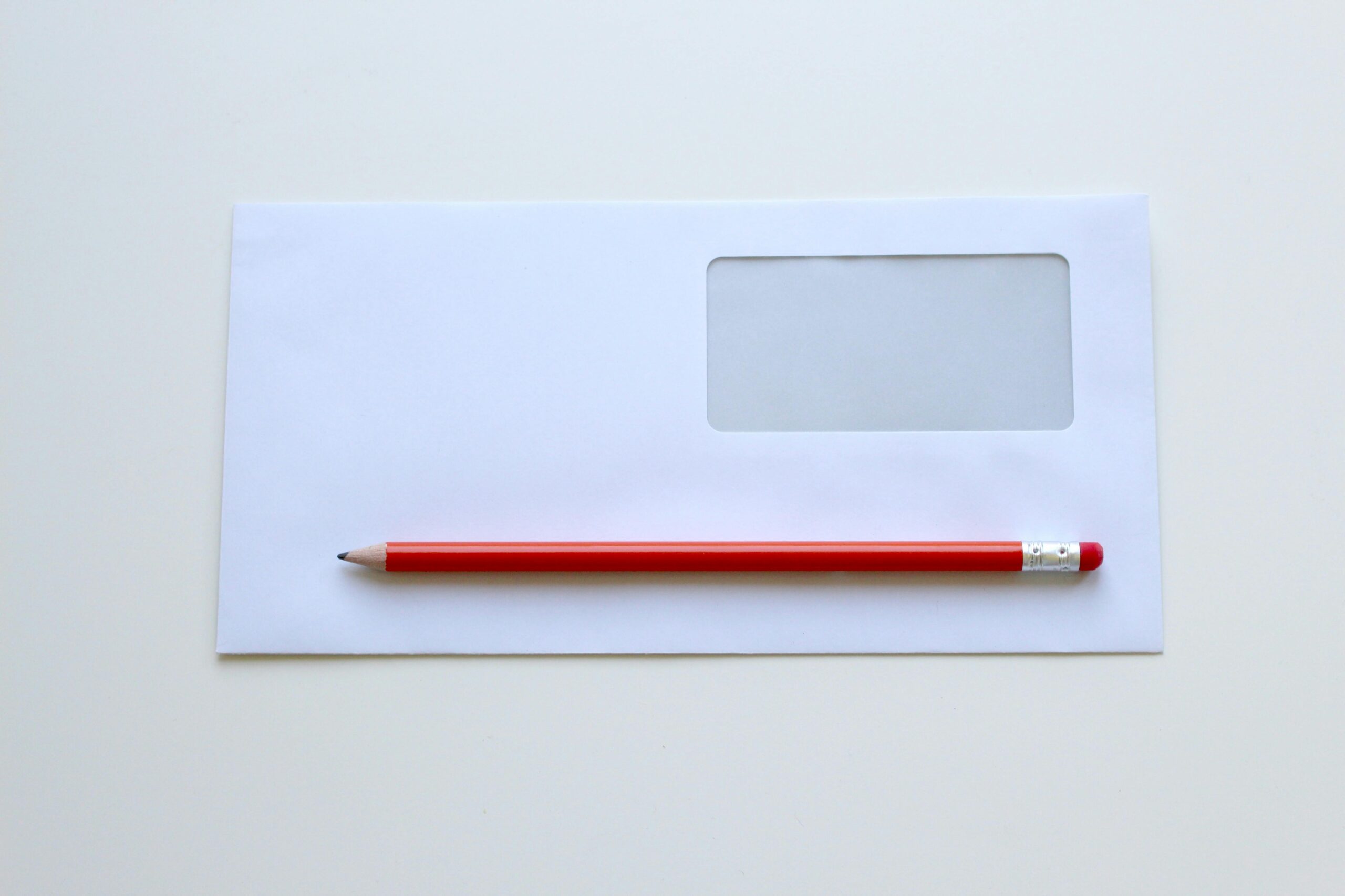 A red pencil lies horizontally on a white envelope with a transparent window.