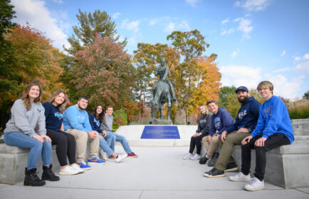 A group of people sitting on a ledge in front of a horseback statue, with trees and a blue sky in the background.