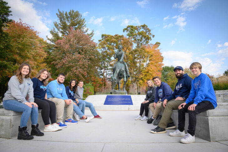 A group of people sitting on a ledge in front of a horseback statue, with trees and a blue sky in the background.