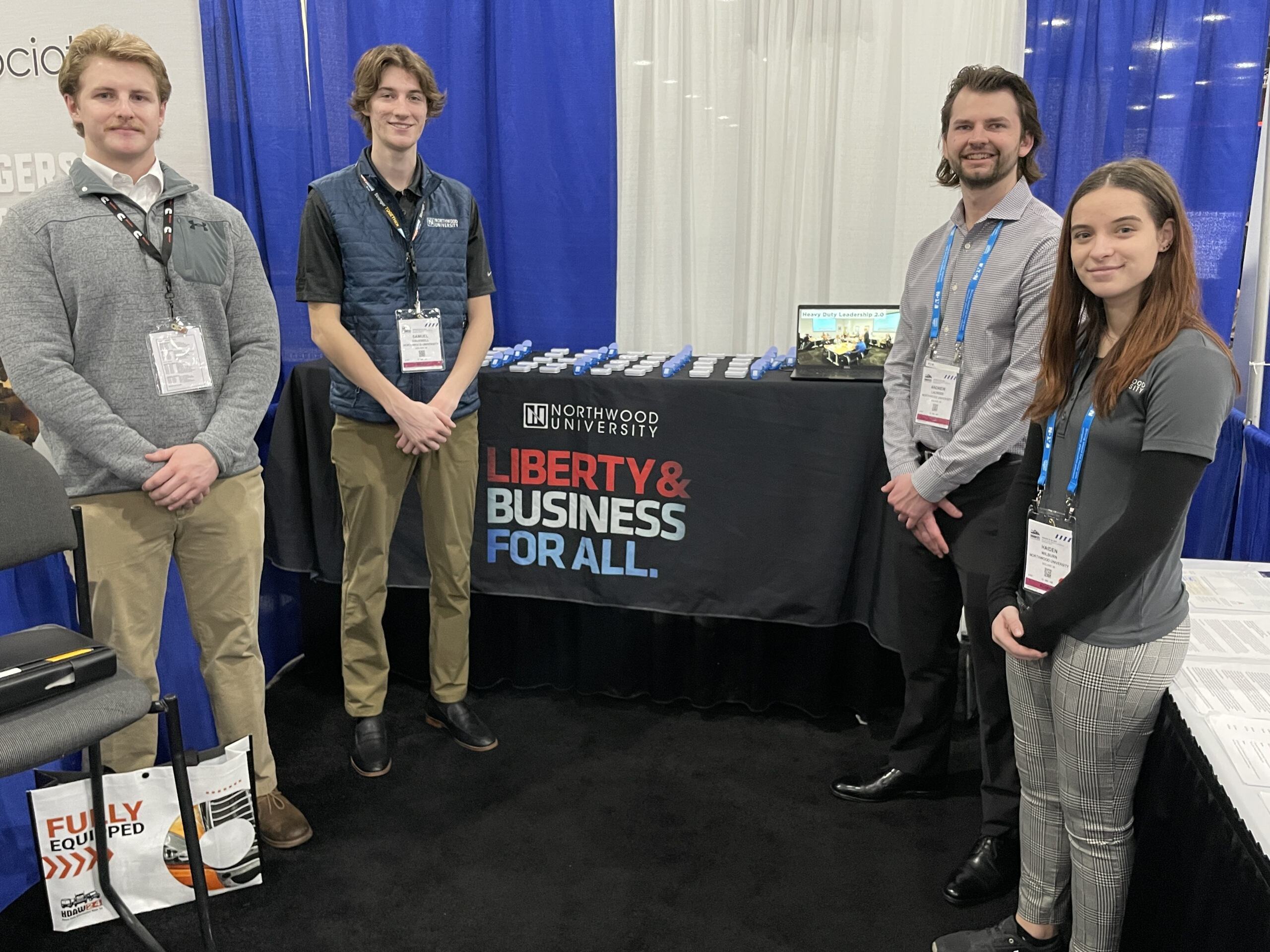 Four people stand in front of a booth with a Northwood University banner at an expo, showcasing the heavy-duty aftermarket industry in North America. They are smiling and wearing conference badges.