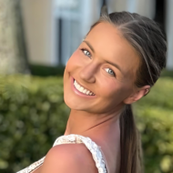 A woman with long hair smiles at the camera, standing outdoors amid lush greenery and a blurred building—capturing a moment as serene as mastering the art of insurance risk management.