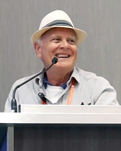 A person wearing a white hat and a light-colored shirt is smiling while embracing the responsibility of addressing the audience at a podium with a microphone.