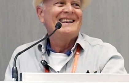 A person wearing a white hat and a light-colored shirt is smiling while embracing the responsibility of addressing the audience at a podium with a microphone.