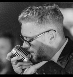 A man speaks or sings into a vintage microphone, elegantly dressed in glasses and a suit, reminiscent of a classic Stafford Dinner scene, all captured in timeless black and white.