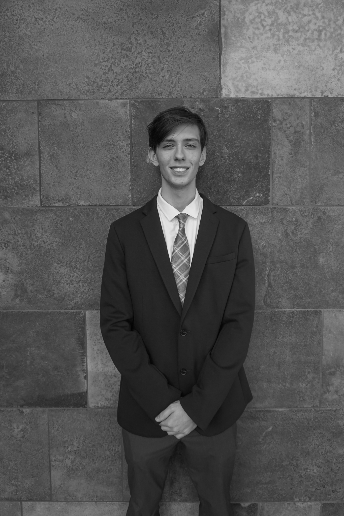 A young man in a suit and tie stands against a textured stone wall, smiling at the camera, reminiscent of an elegant Stafford dinner gathering. Black and white photo.