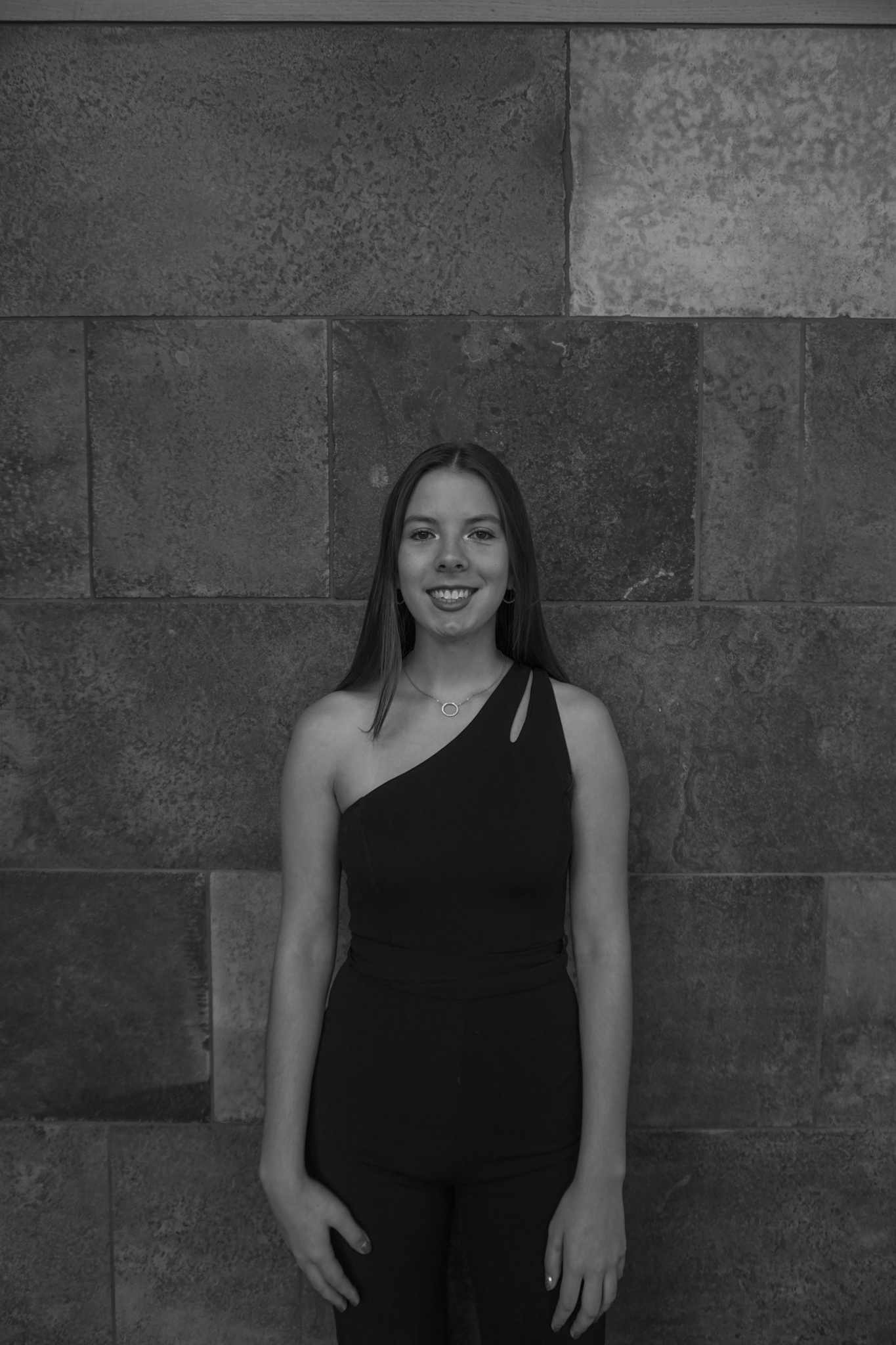 Woman in a chic one-shoulder top stands against a textured stone wall, reminiscent of the sophisticated elegance of a Stafford dinner setting.