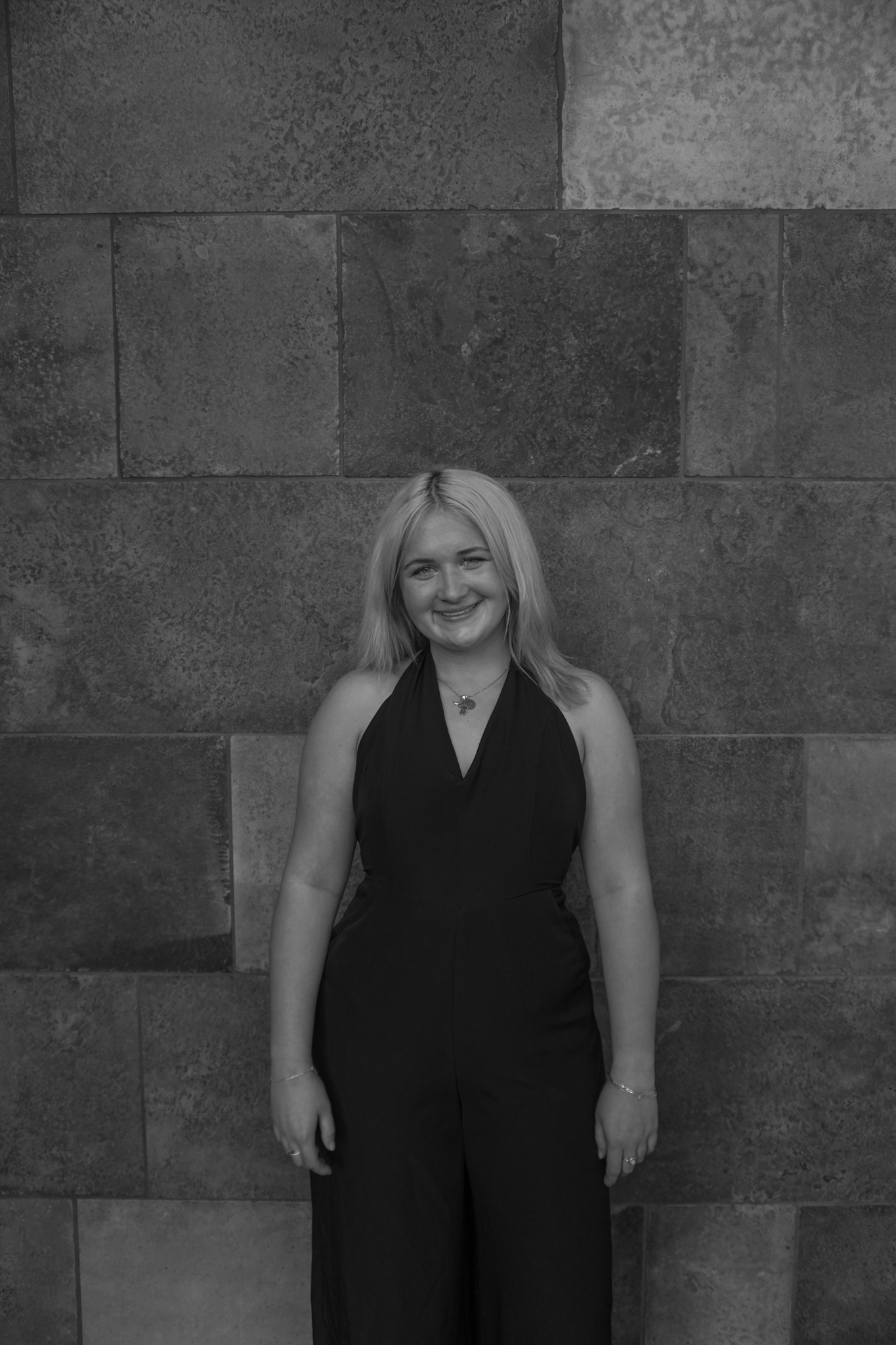 A person with long hair, wearing a sleeveless outfit, stands gracefully in front of a textured stone wall, reminiscent of an evening at Stafford Dinner.