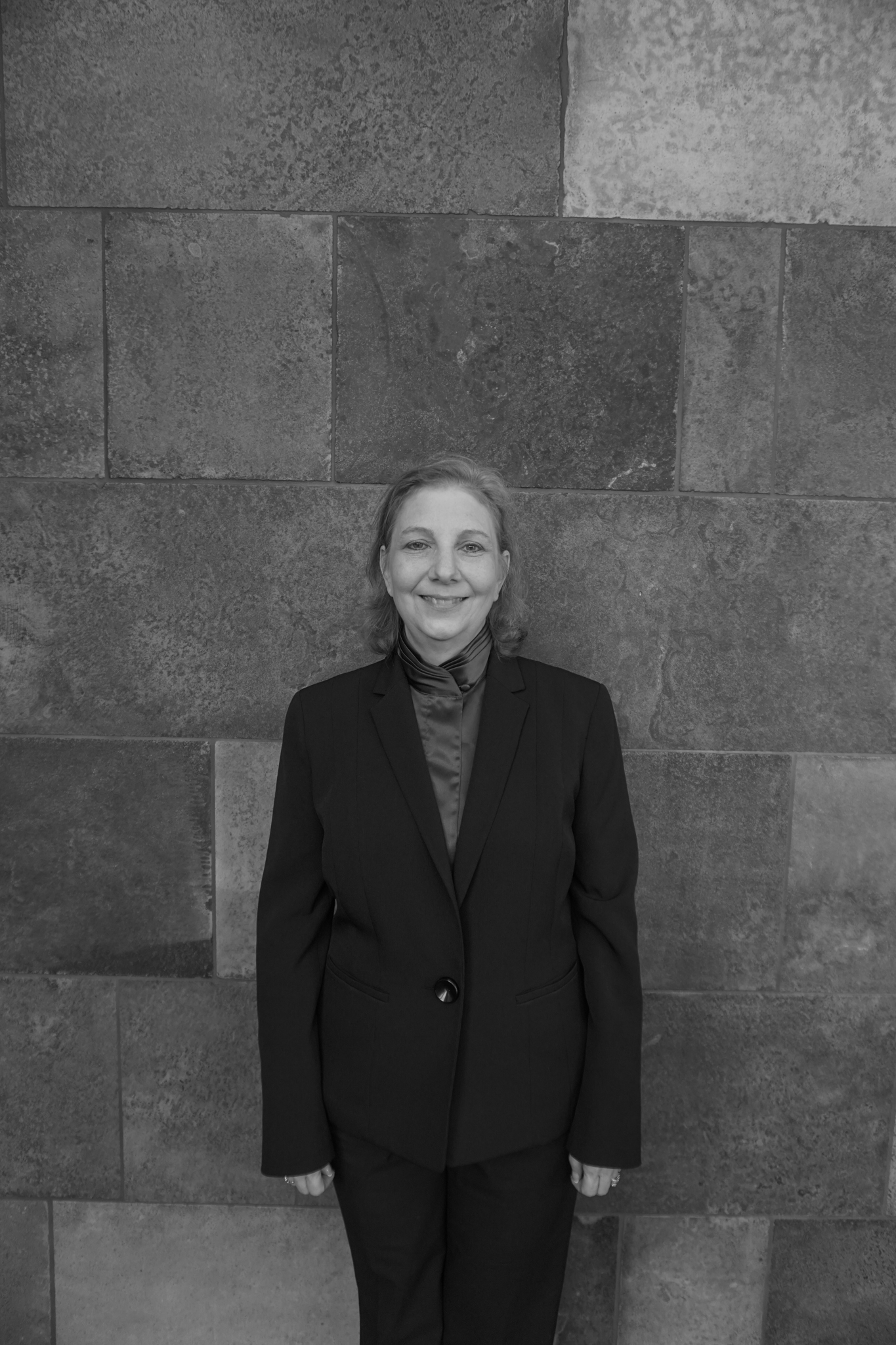 A person in a suit, perhaps heading to the Stafford Dinner, stands against a textured stone wall, smiling warmly at the camera. The image captures this timeless moment in elegant black and white.