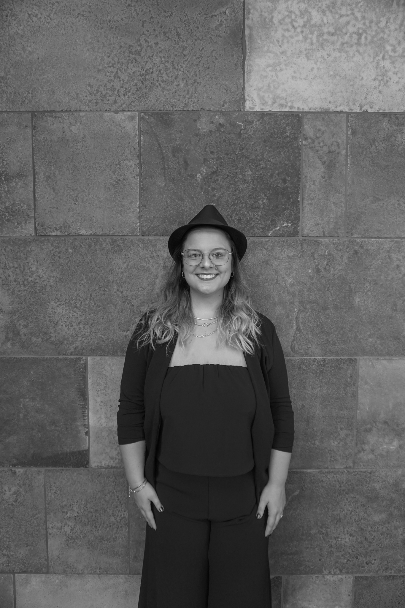 A person wearing glasses and a hat stands smiling against a stone wall, reminiscent of a stylish evening ready for a Stafford dinner. Black and white photo.