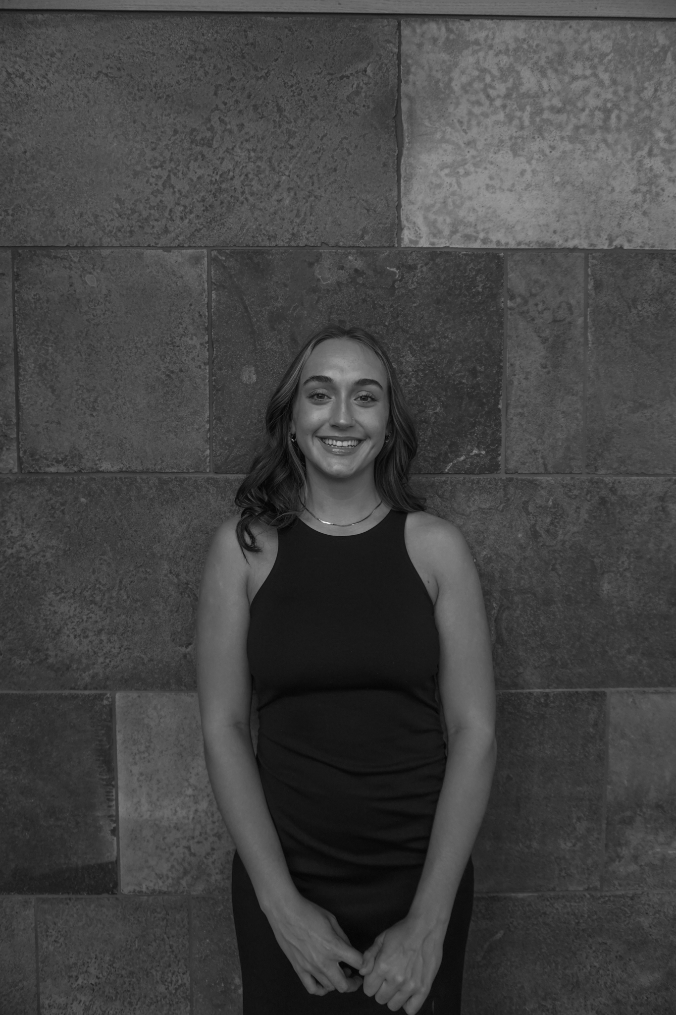 A woman in a sleeveless dress stands smiling in front of a textured stone wall, reminiscent of vintage Stafford Dinner gatherings. The image is captured in elegant black and white.