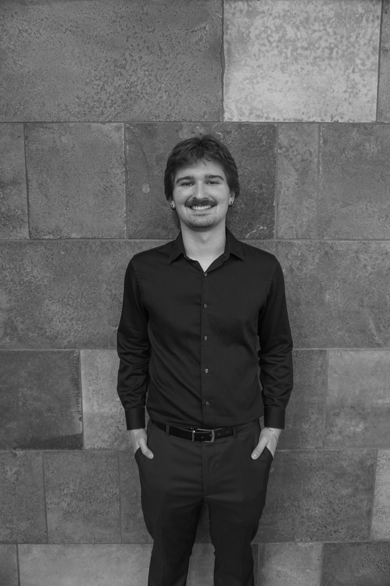 Black and white photo of a person with a mustache, wearing a dark shirt and trousers, standing against a tiled wall with an air of Stafford Dinner sophistication, hands casually tucked in pockets.